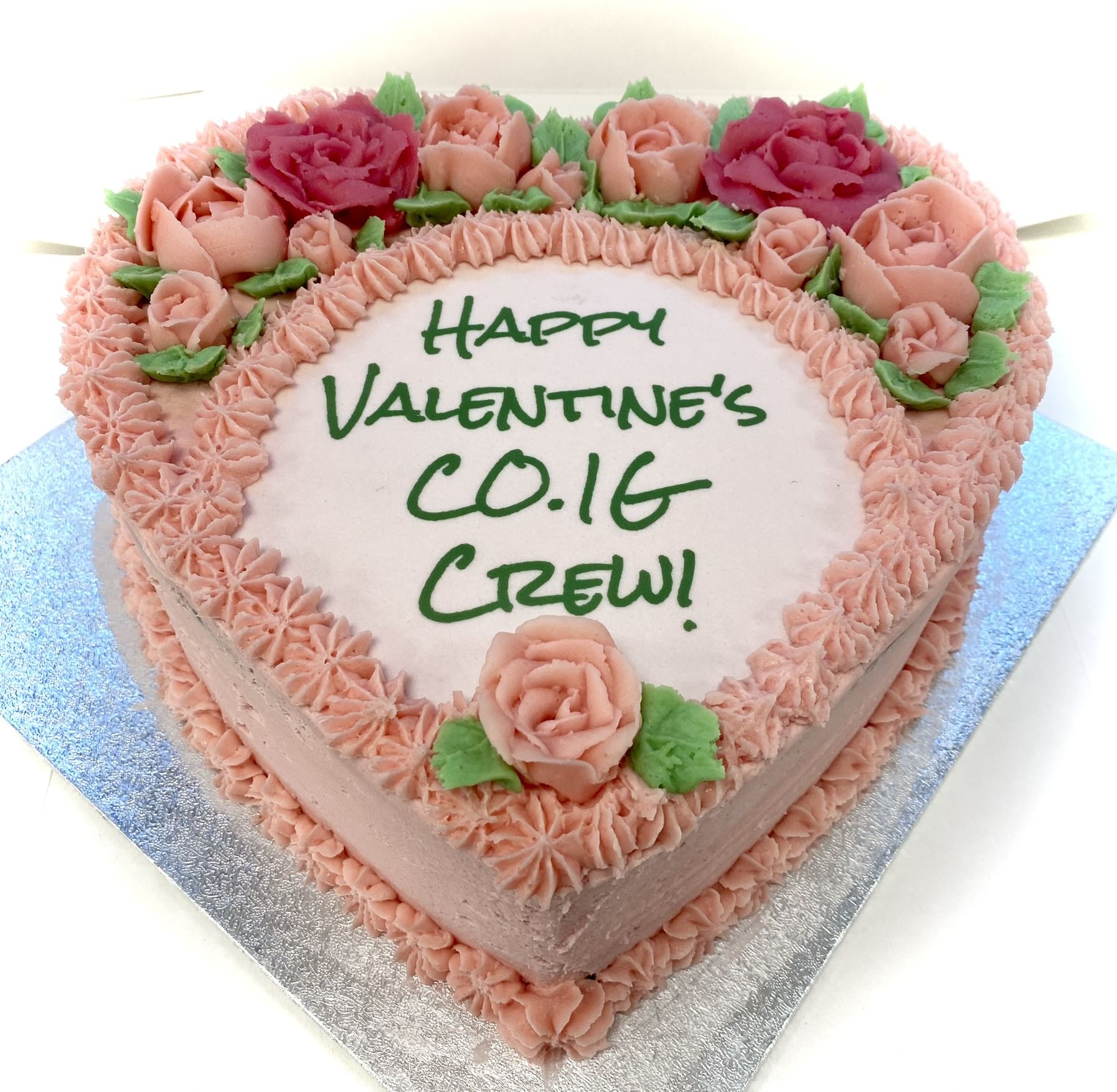 A heart-shaped cake with pink frosting, decorated with pink and red roses, and green leaves. The cake has the message 'Happy Valentine's CO.IG Crew!' written in green icing in the center. This special gift of love is placed on a silver square board.